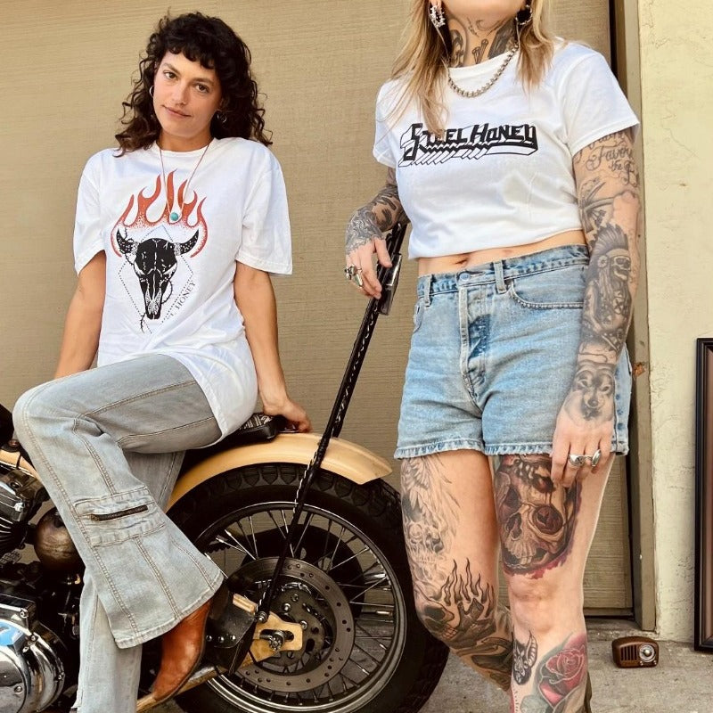 two women modeling steel honey shirts with steel honey's chopper. one model is sitting on the seat and the other is standing