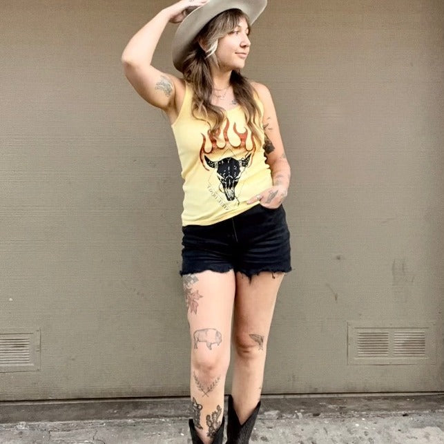 Model wearing a yellow tank top, with steel honey lettering in black across the front in a stylized font. Model is wearing black jean cutoffs, black boots and western style hat